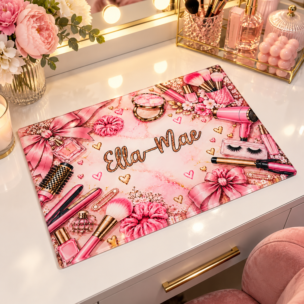 A personalized pink makeup desk mat featuring the name 'Ella-Mae' in gold glitter script, surrounded by illustrated makeup items, bows, flowers, and hearts, displayed on a white vanity desk.