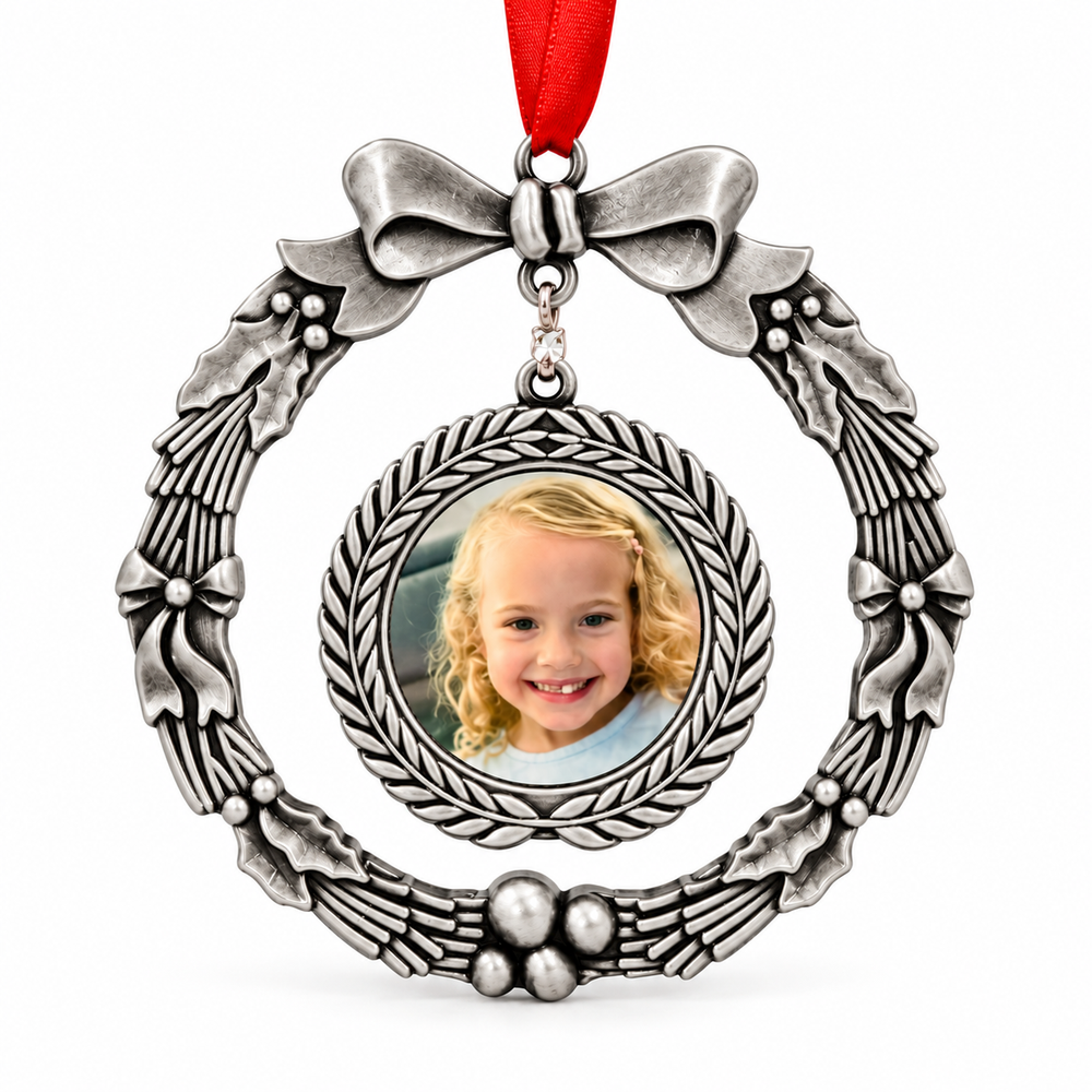 A silver-toned metal Christmas wreath photo ornament with a red hanging ribbon and a circular inset frame showing a young girl's portrait, photographed on a white background.