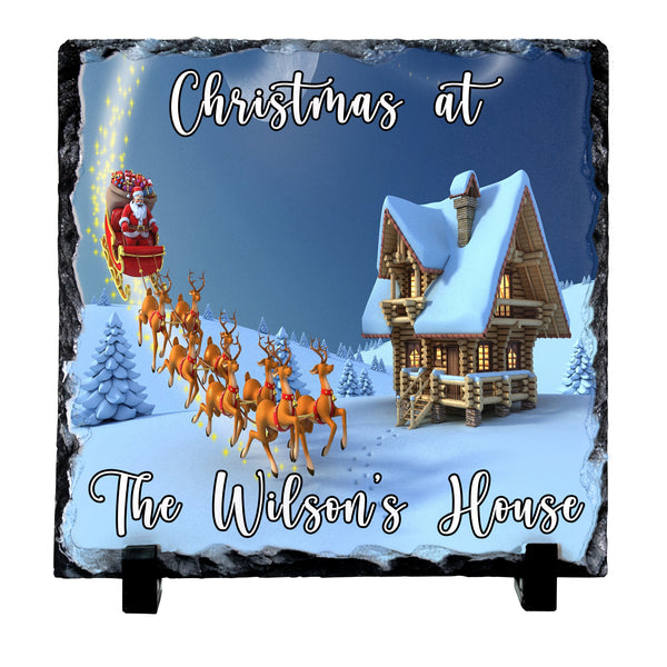 Personalised slate Christmas plaque featuring a snowy cabin, Santa in his sleigh, and the text 'Christmas at The Wilson's House' on a dark slate background.