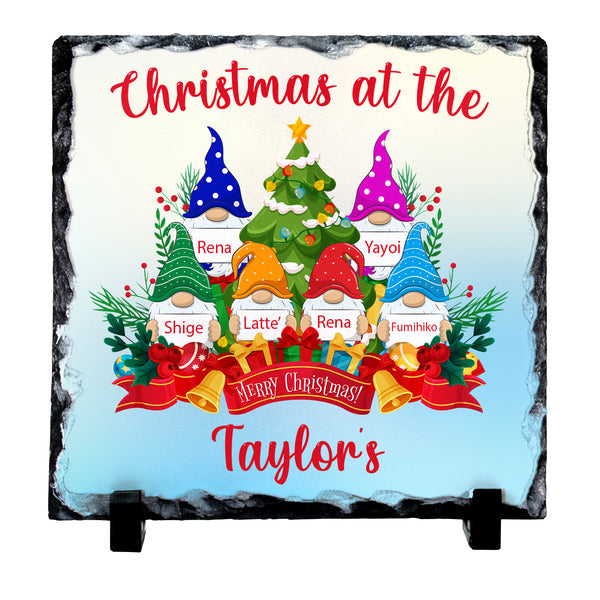 Personalised square slate Christmas plaque on a black display stand, featuring a festive illustration of gnome characters around a decorated Christmas tree with customizable names printed on the design.
