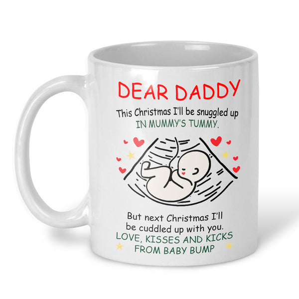 A white ceramic mug with a printed Christmas-themed message for a baby shower. The text reads 'DEAR DADDY' in red, followed by black and green text, a simple line drawing of a baby inside an ultrasound scan outline with small red and yellow hearts, and more text below.