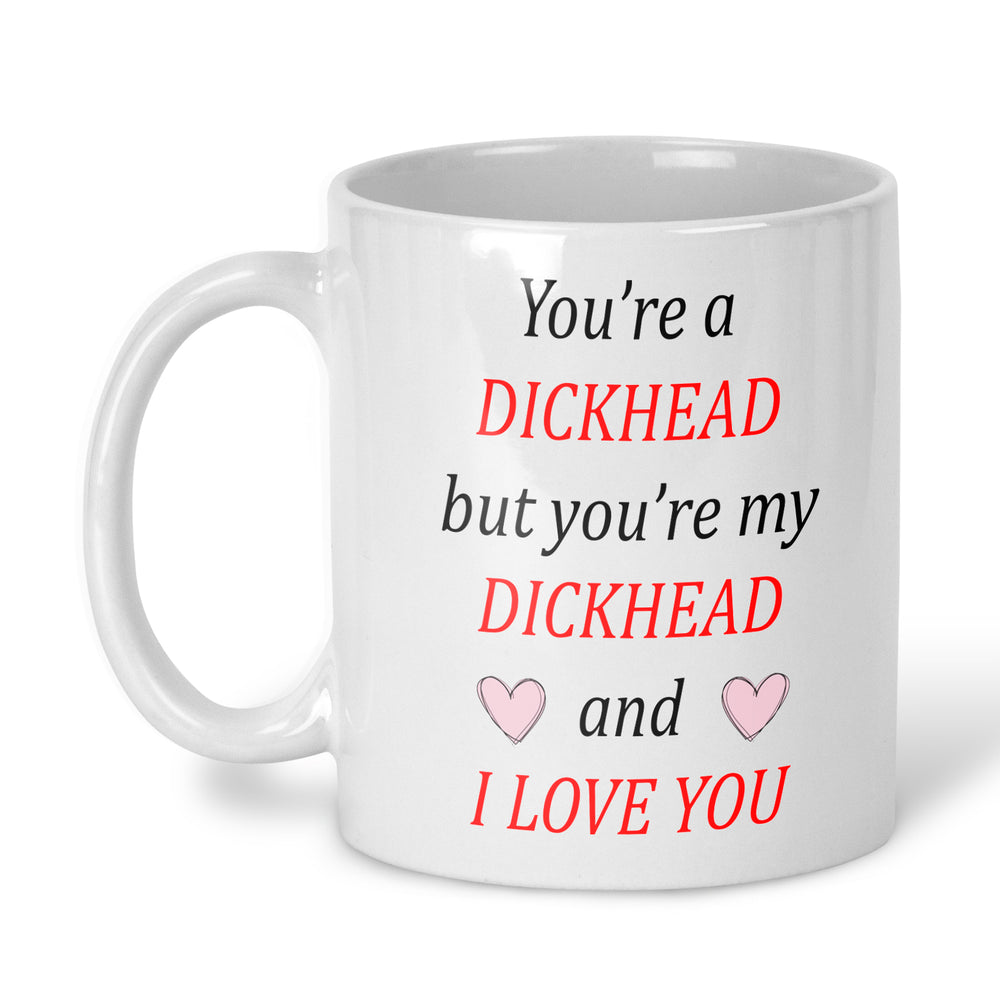 A white ceramic mug with a humorous printed message on the side. The text reads 'You're a DICKHEAD but you're my DICKHEAD I LOVE YOU' in black and red lettering, accented with small pink hearts.