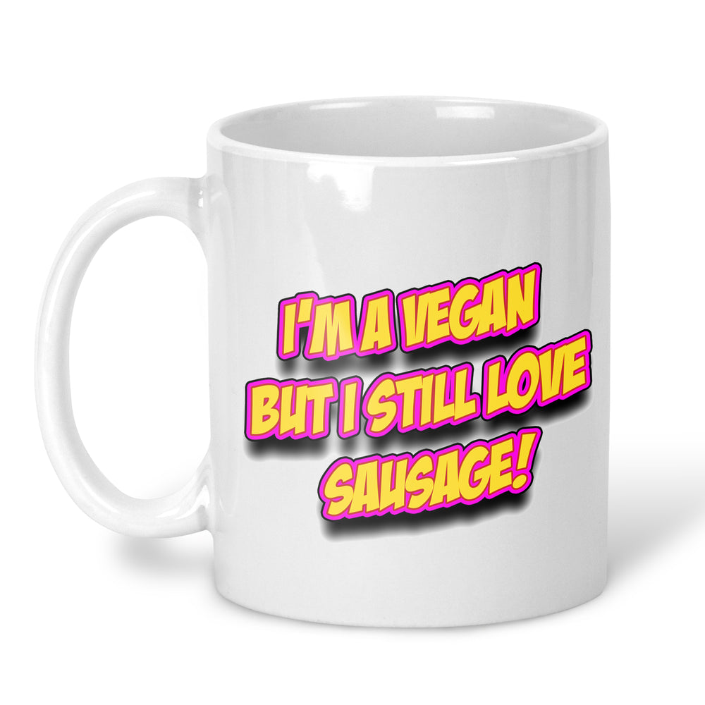 A white ceramic mug with a bold graphic text design that reads 'I'M A VEGAN BUT I STILL LOVE SAUSAGE!' in yellow letters with a pink outline and black shadow, set against a plain white background.
